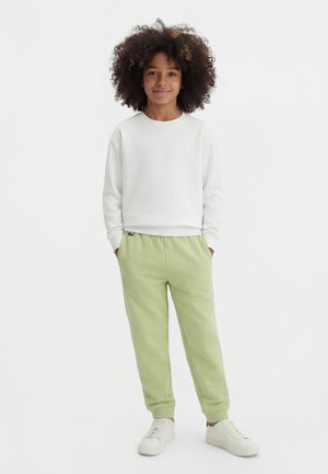 Kind met krullend haar draagt een witte trui, lichtgroene joggingbroek en witte sneakers, staat met handen in de zakken.