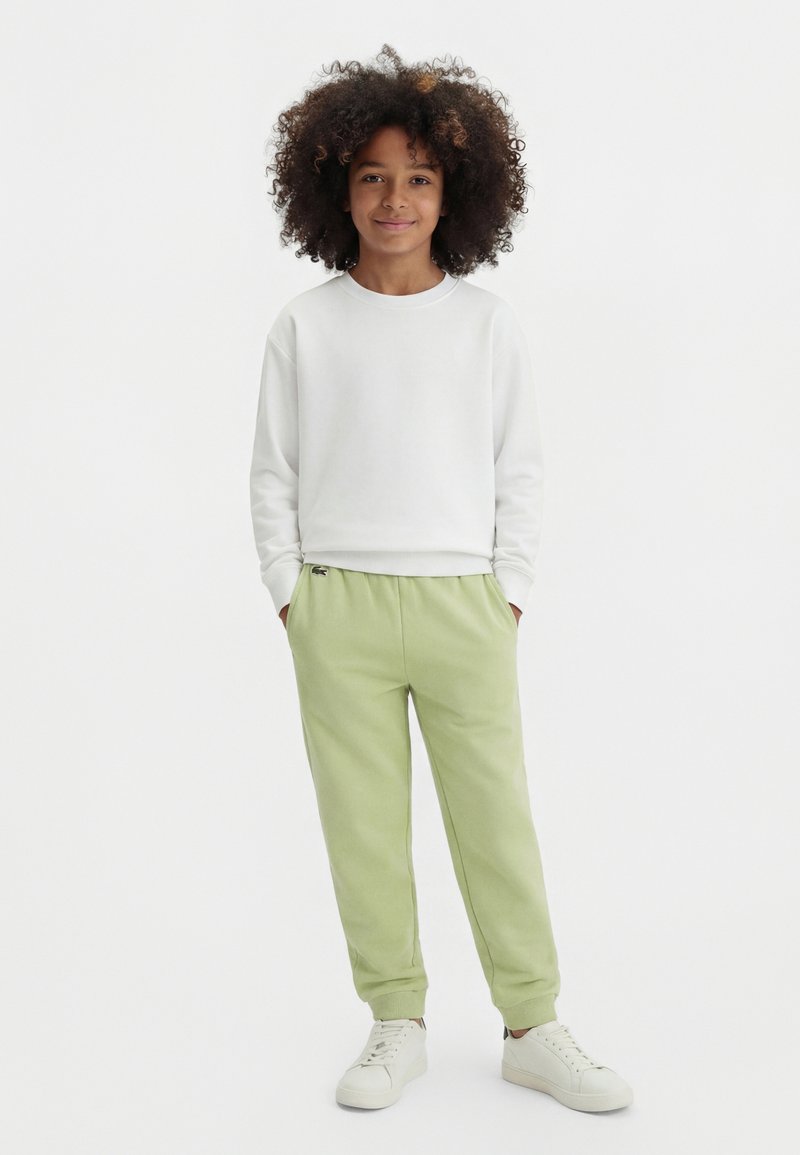 Παιδί με κατσαρά μαλλιά που φοράει λευκό φούτερ, ανοιχτό πράσινο παντελόνι τζόγκινγκ και λευκά αθλητικά παπούτσια, στέκεται με τα χέρια στις τσέπες.