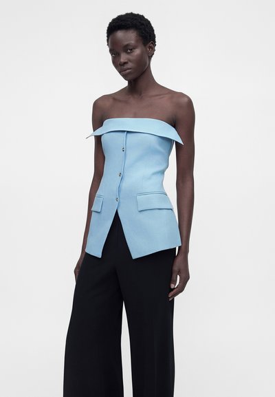 Γυναίκα με αμάνικο ανοιχτό μπλε τοπ με κουμπιά και τσέπες και μαύρο παντελόνι με φαρδιά πόδια, στέκεται μπροστά σε απλό φόντο.