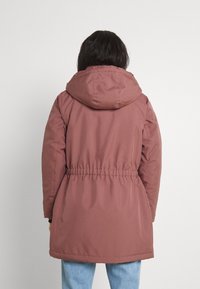 Veste longue à capuche mauve avec taille cintrée et détails élastiques. Texture douce, design simple et silhouette légèrement évasée.