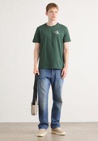 Grøn bomulds crew-neck t-shirt med et hvidt logo, parret med lyseblå denim jeans og beige sneakers, mens der holdes en lille grå taske.