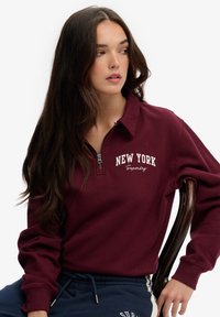 Burgundowa bluza z zamkiem błyskawicznym z napisem i logo "NEW YORK" w białym kolorze. Miękka tkanina z prążkowanymi mankietami, w stylu casual z granatowymi spodniami.