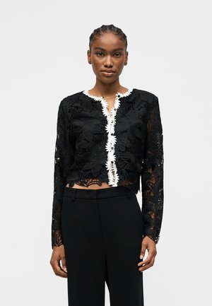 Γυναίκα που φορά μαύρο μακρυμάνικο σακάκι με λουλουδένια δαντέλα και άσπρο περιθώριο κατά μήκος του εμπρός ανοίγματος, σε συνδυασμό με μαύρο παντελόνι ψηλόμεσο.