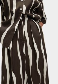Chemise boutonnée à rayures verticales marron et blanches avec un pantalon large plissé assorti, main dans la poche.