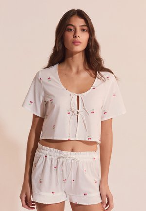 Junge Frau trägt ein weißes, kurzärmeliges Crop-Top und passende Shorts mit kleinen roten Kirschmotiven,steht vor einem einfarbigen beigen Hintergrund.