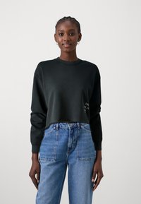 Sudadera negra recortada con mangas largas, escote redondo y un dobladillo desgastado. Combinada con jeans azules de talle alto con bolsillos frontales.