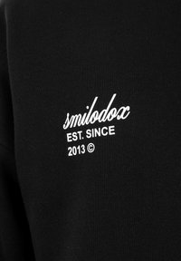 Zwarte stof met witte tekst "smilodox EST. SINCE 2013 ©" erop gedrukt, waarschijnlijk op een kledingstuk.