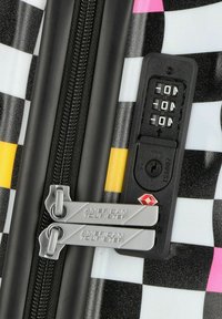 Valigia rigida a scacchi bianco e nero con dettagli gialli e rosa, dotata di doppi ganci in metallo per la zip e di una combinazione per la chiusura.