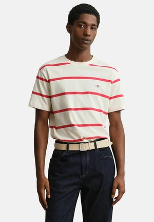 Junger Mann, der ein cremefarbenes T-Shirt mit roten horizontalen Streifen, dunkelblaue Jeans und einen hellbeigen Gürtel vor einfarbigem Hintergrund trägt.