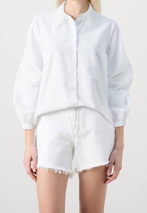 Femme portant un chemisier blanc boutonné avec col et poignets en dentelle, assorti à un short en jean blanc effiloché, sur un fond uni.