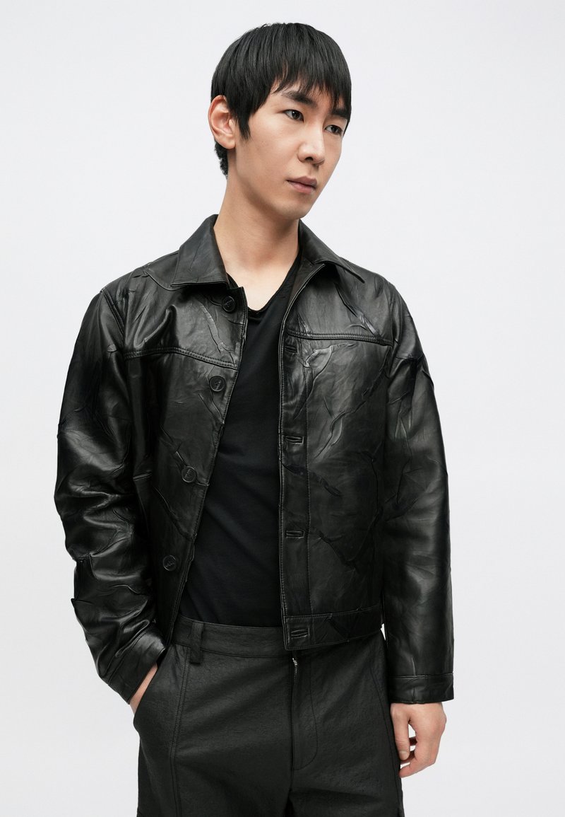 Veste en cuir noire à surface texturée, dotée d'une fermeture à boutons, d'un large col et d'une coupe courte. Portée sur une chemise noire.