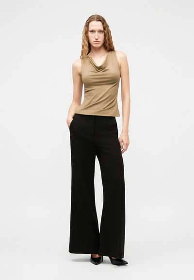 Γυναίκα με μακριά σγουρά μαλλιά που φοράει αμάνικο καφέ τοπ και μαύρο παντελόνι ψηλόμεσο με φαρδιά μπατζάκια, όρθια με το ένα χέρι στην τσέπη.