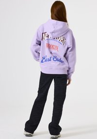 Persona con cabello rojo largo que lleva una sudadera lila con el texto Nueva York, Brooklyn y East Side, pantalones negros y zapatillas blancas, de espaldas.