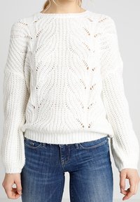 Femme portant un pull en grosse maille blanche avec un motif texturé, associé à un jean en denim bleu, debout devant un fond uni.