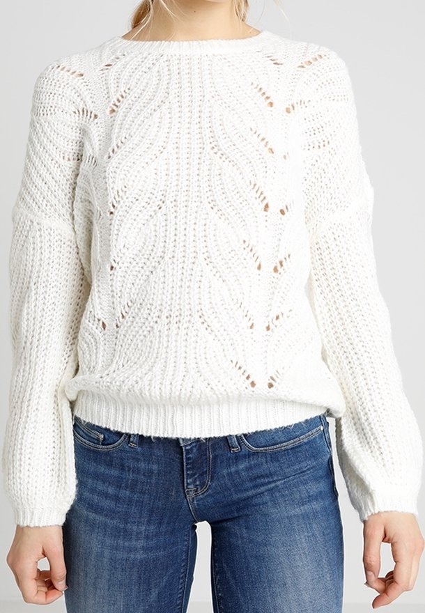 Femme portant un pull en grosse maille blanche avec un motif texturé, associé à un jean en denim bleu, debout devant un fond uni.