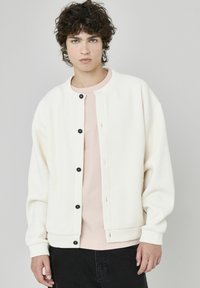 Veste blanche à manches longues à côtes, avec des boutons-pression noirs, portée par-dessus une chemise rose pâle. La veste a un col rond et une coupe décontractée.