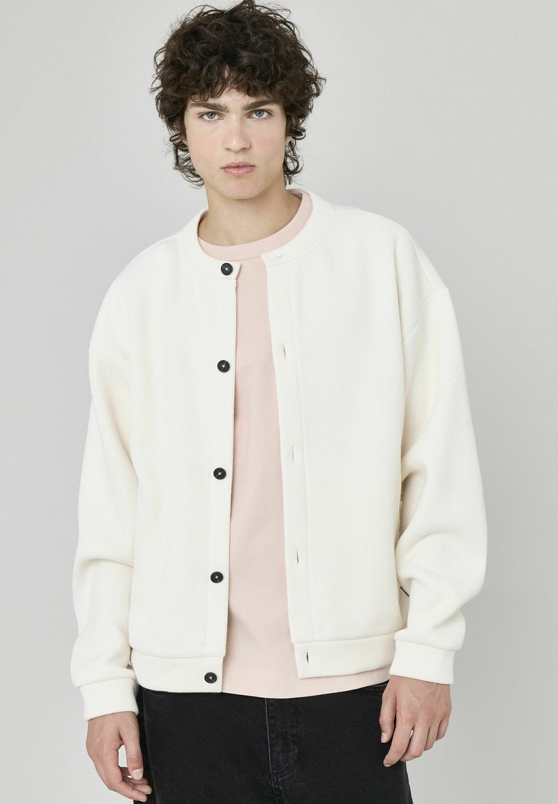 Veste blanche à manches longues à côtes, avec des boutons-pression noirs, portée par-dessus une chemise rose pâle. La veste a un col rond et une coupe décontractée.