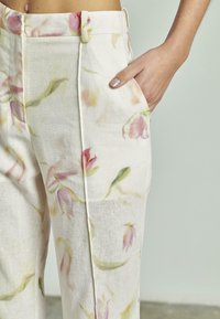Close-up van een persoon in beige broek met zachte roze en groene bloemmotieven, hand rustend in de zijzak tegen een effen achtergrond.