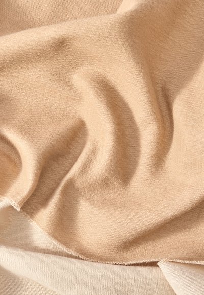 Μαλακό μπεζ ύφασμα με ομαλή υφή, πτυχωμένο για να δείχνει απαλά πτυχώσεις και ζάρες σε ένα ανοιχτό μπεζ φόντο.