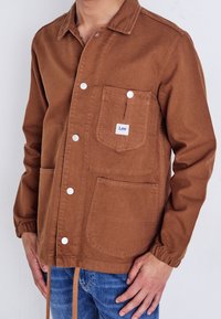 Man som bär en brun knäppt jacka med bröstficka märkt "Lee" och elastiska ärmmuddar, i kombination med blå jeans, står mot en vit bakgrund.