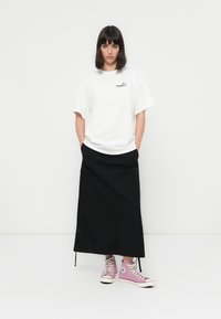 Mujer de pie frente a un fondo blanco, vestida con una camiseta gráfica blanca, una falda larga negra con bolsillos y zapatillas de deporte de caña alta rosas.