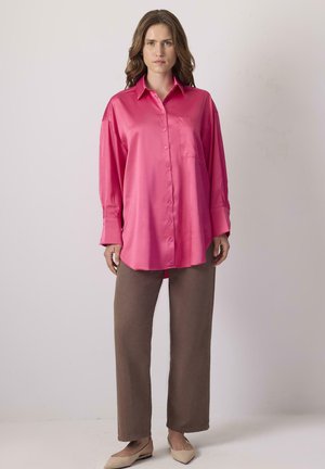 Femme portant une chemise ample rose vif à boutons, un pantalon marron à jambes droites et des ballerines beige à bouts pointus, debout devant un fond clair uni.
