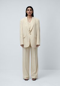 Kremni suit s pinstripi z ohlapnim krojem, ki vključuje blazer z enim gumbom in široke hlače. Gladka tekstura, minimalno detajliran, brez vidne kovinske opreme.