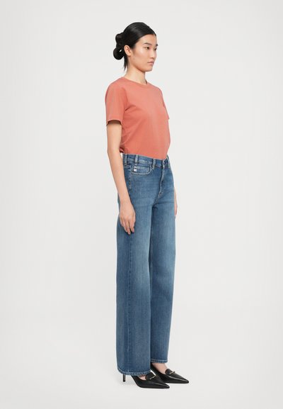 Mulher de pé de lado, vestindo uma t-shirt cor ferrugem, umas calças de ganga largas de cintura alta azuis e sapatos de salto agudo pretos, contra um fundo branco.