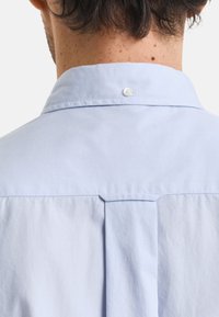 Hellblaues Hemd mit kleinem Kragen und Knopfverschluss sowie einer mittigen Falte auf der Rückseite, aus einem glatten Stoff gefertigt.