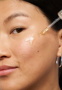 Serum in einem Tropfer, das auf die Haut aufgetragen wird; klar und leicht viskos, mit einem goldenen Schimmer, auf einem glatten, strahlenden Teint. Eine silberne Creolen-Ohrringe ist sichtbar.