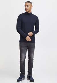 Maglione a collo alto blu navy con maniche lunghe abbinato a jeans slim-fit grigi e scarpe sportive blu navy. Tessuto morbido, design aderente.