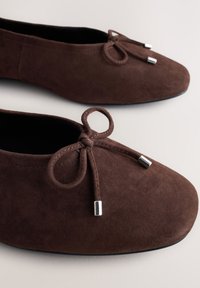 Ballerines en daim marron avec un petit nœud et des embouts métalliques argentés sur les lacets, photographiées sur une surface claire.