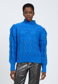 Maglione blu lavorato a maglia con collo alto, caratterizzato da motivi floreali e fogliari, polsini e orlo a coste, e una finitura testurizzata. Abbinato a pantaloni metallici argento.