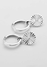 Silverörhängen i form av hoopar med en cylindrisk berlock med strålande linjer och en central ädelsten, som har en polerad yta och en slät textur.