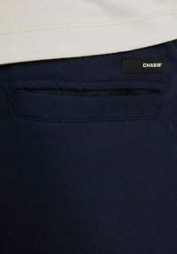 Námornícke modré nohavice s zadnou kapsou a čiernou nálepkou s nápisom "CHASIN'." Látka má hladký povrch a jemný diagonálny vzor.