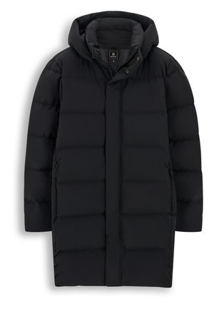 Un homme pose vêtu d’un long manteau matelassé noir, d’un bonnet et de gants noirs, d’un pantalon foncé et de baskets noires sur un fond blanc uni.