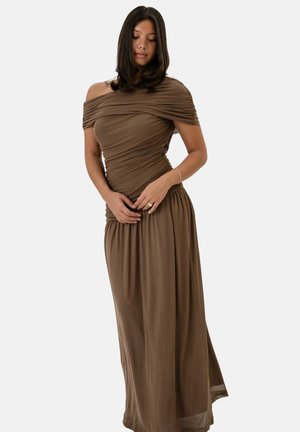 Mujer con cabello oscuro que lleva un vestido largo marrón sin hombros con tela fruncida, de pie frente a un fondo liso.