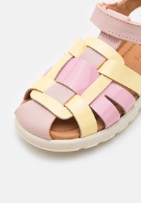 Värviline sandal, millel on roosa ja kollane läikiv kootud rihm, avatud varvaste disain ning pehme pruun sisesole ja valge välistald.
