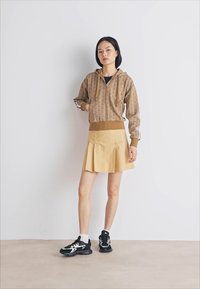 Bruine hoodie met patroon en rits, gecombineerd met een beigekleurige plooirok en zwarte sportschoenen met witte accenten.