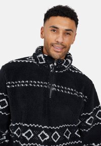Pullover in pile nero con motivi geometrici bianchi, colletto con zip a metà e superficie morbida e strutturata. Design caldo e accogliente.