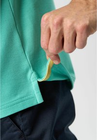 Turkos kortärmad polo skjorta i texturerad tyg, med en gul etikett i fållen, bärs över mörka byxor. Hand som håller i tygkanten.