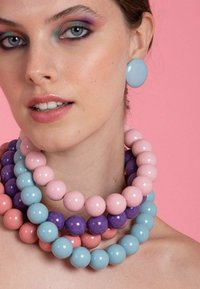 Nainen, jolla on rohkeat pastellivärit silmämeikissä, käyttää suuria sinisiä pyöreitä korviksia ja monia paksuja helminauhoja vaaleanpunaisessa, violetissa ja sinisessä värissä vaaleanpunaisen taustan edessä.
