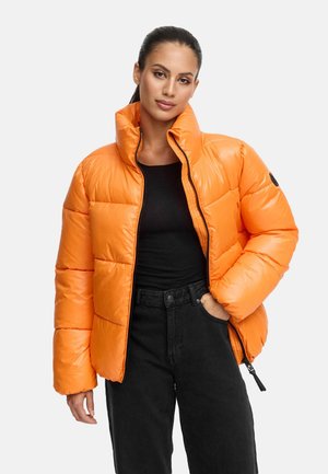 Giubbotto imbottito di un vivace arancione con colletto alto, design trapuntato e dettagli in zip neri. Abbinato a una maglietta nera e jeans scuri.
