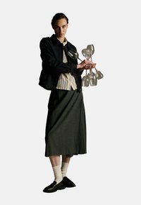 Modèle portant une veste noire par-dessus une chemise rayée et une jupe plissée grise, tenant un groupe de sept verres en verre transparent. Chaussures noires et chaussettes crème.