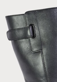 Acercamiento a una bota de cuero negra que muestra una costura cosida y una correa decorativa con un bucle de metal cerca de la parte superior.