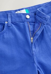 Pantalon bleu coupe droite avec fermeture éclair argentée partiellement ouverte et bouton métallique supérieur visible à la taille sur fond blanc.