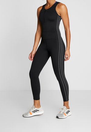 Kvinde iført en tætsiddende sort atletisk jumpsuit med grå striber, parret med grå og orange løbesko, stående foran en neutral baggrund.