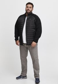 Black insulated jacket with a quilted front, grey long pants, and navy athletic shoes. Styling: zip-up with side pockets and contrasting sleeves.