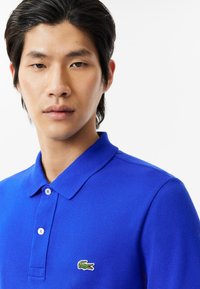 Blå pikétröja i texturerat tyg, med en treknappsslutning och en grön broderad krokodil-logotyp på vänster bröst.