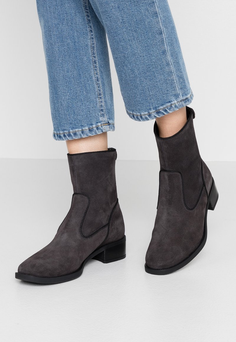 Botas de tobillo de ante en gris oscuro con punta cuadrada, tacón bajo y detalles de costura negra. Combinadas con pantalones de mezclilla azul recortados.